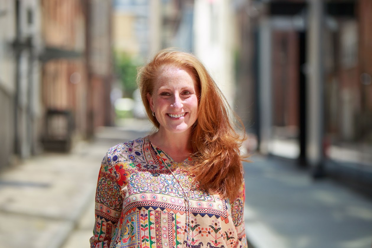A smiling woman with long red hair stands in an urban alleyway, wearing a colorful patterned blouse.